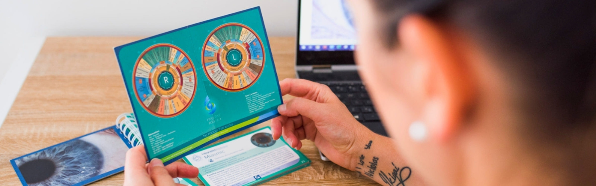 Diane Hackner holding an iridology chart with a laptop on a wooden table.