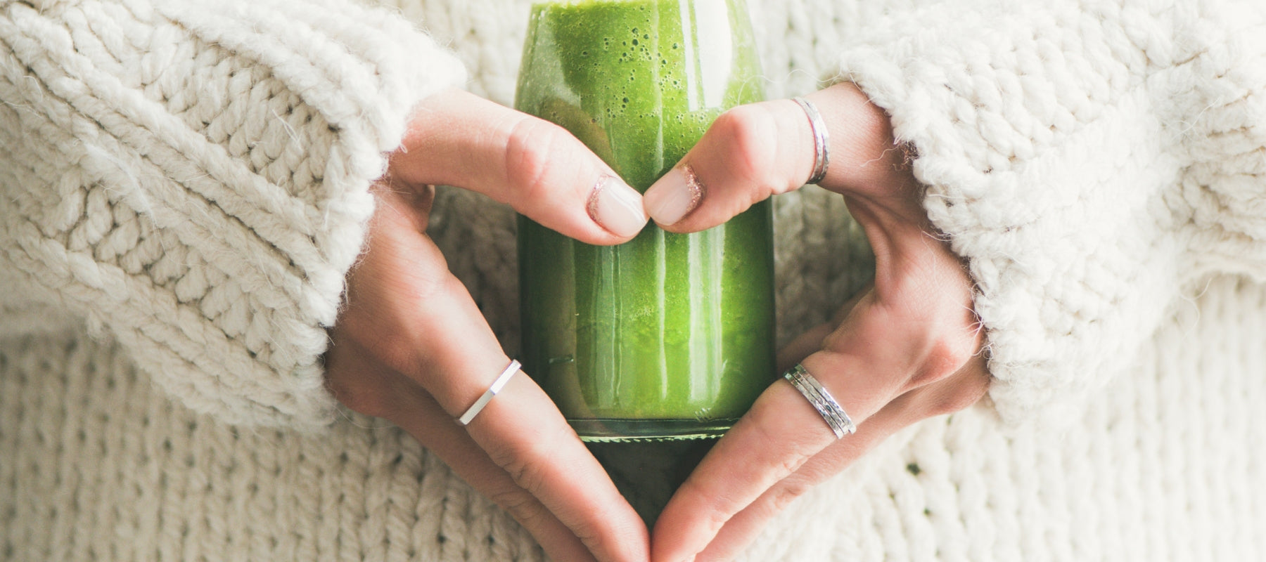 Person holding a green smoothie in a heart shape with a white textured background. Self led detox protocols.
