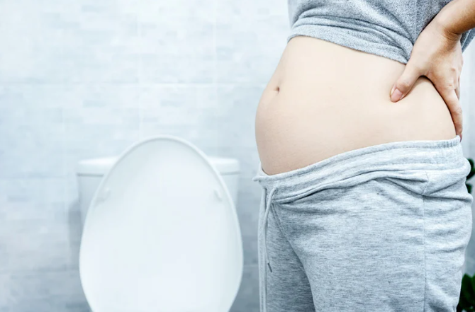 Person wearing grey sweatpants standing in front of a toilet showing how bloated they are.